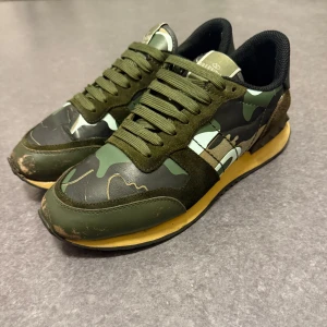 Camouflage sneakers med nitar - Snygga valentino rock runners i  camouflagefärg med gröna och svarta toner. Skorna har en unik design med nitar på hälen och snörning framtill. Tillverkade i en kombination av mocka och syntetmaterial, vilket ger en cool och modern look.  Skorna är i storlek 41 men passar 42