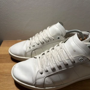  Saint Laurent skor - Snygga vita sneakers från Saint Laurent med unika läppdetaljer i blått och rött på hälen. Skorna har klassisk snörning och är tillverkade i skinn. Skicket är ungefär 7/10 och har fortf mycket at ge! Kom med prisförslag och strl är 42! 