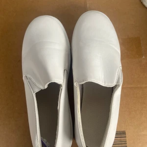 Vita slip-on sneakers - Säljer ett par vita slip-on sneakers i syntetmaterial. De har en enkel och stilren design utan snörning, vilket gör dem lätta att ta på och av. Perfekta för en avslappnad stil.