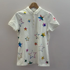 Vit pikétröja med stjärnor från U.S. Polo Assn. - Säljer en vit pikétröja från U.S. Polo Assn. med färgglada stjärnor i olika storlekar. Tröjan har korta ärmar och en klassisk krage med knappar framtill. Perfekt för en färgglad och lekfull stil!