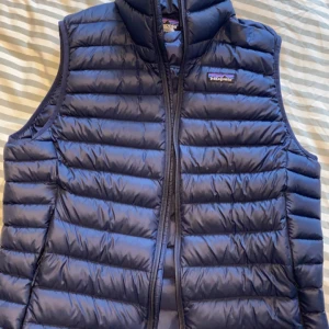 Blå dunväst från Patagonia - Säljer en blå dunväst från Patagonia med dragkedja. Västen är quiltad och perfekt för kyligare dagar. Den har en klassisk design med en liten logga på bröstet. Finns ett litet hål under ärmen går att fixa med lagnings lappar. 