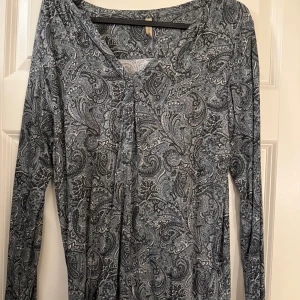 Mönstrad långärmad topp från Soyaconcept - Snygg långärmad topp från Soyaconcept med ett detaljerat paisleymönster i grå och svarta nyanser. Perfekt för en stilren look. Toppen har en v-ringad hals och är tillverkad i ett mjukt material.