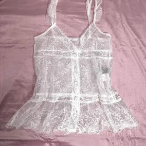 Vit spetslinne - *japanska märker*.                                                                    *det är genomskinligt*Säljer ett superfint vitt spetslinne med v-ringning och volangdetaljer. Det är ärmlöst och har en luftig passform med knappar framtill. 