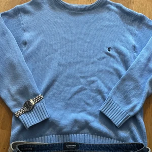 Ralph Lauren Vintage Stickad Tröja Ljusblå - Säljer en stilren ljusblå tröja från Ralph Lauren i 100% bomull. Tröjan har en liten broderad logotyp på bröstet och ribbade muddar vid ärmslut och nederkant. Perfekt för en avslappnad och snygg look. Enda deffekten är att storkleks kappen slitits av, men är ändå säker på atta storleken är XL.