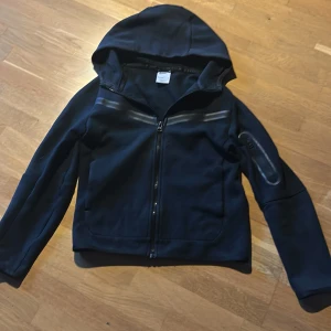 Svart Nike tech fleece - Snygg svart Nike tech fleece från Nike med huva och dragkedja framtill. Jackan har långa ärmar och en stilren design med diskreta detaljer. Perfekt för en sportig look. 137-147 Cm