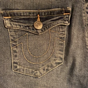 True Religion Jeans - Snygga blå jeans från True Religion med broderade detaljer på bakfickorna. De har en klassisk bootcut-stil. Köpte här på plick men säljer då det ej passade. Innerbenslängden är cirka 96 och midjemått w28.