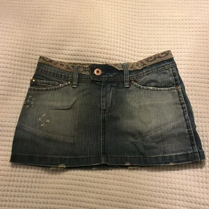 Jeanskjol från Suf Denim - Lågmidjad Snygg jeanskjol från Suf Denim med spetsdetaljer i midjan och coola korsstygnsbroderier på fickorna. Perfekt för en avslappnad stil. Kjolen har en klassisk femficksdesign och en knapp framtill. 