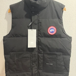 Svart dunväst från Canada Goose - Säljer en svart dunväst från Canada Goose med broderad logotyp på bröstet. Västen har praktiska fickor och knappar framtill. Perfekt för kyliga dagar. Storlek L, passar M också
