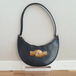 Svart axelväska med gulddetalj från Zara - Snygg svart axelväska från Zara med en unik guldfärgad detalj på framsidan, i nyskick. Väskan har en dragkedja upptill och en enkel axelrem. Perfekt för att ge en touch av elegans till din outfit. 25×13cm, hand 45cm