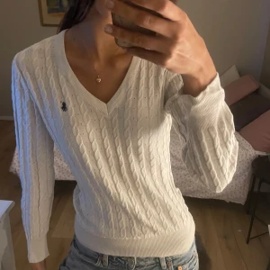 Vit kabelstickad tröja från Ralph Lauren - Säljer en stilren vit kabelstickad tröja från Ralph Lauren med v-ringning och det klassiska logotypbroderiet på bröstet. Tröjan har långa ärmar och ribbade muddar vid ärmslut och nederkant. Perfekt för en elegant och avslappnad look.