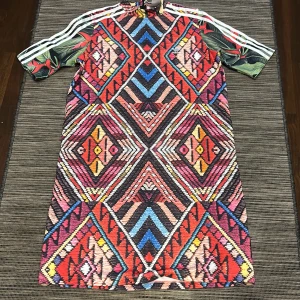 Färgglad mönstrad klänning från Adidas - Säljer en unik och färgglad klänning från Adidas med ett geometriskt mönster i rött, blått och gult. Klänningen har korta ärmar med Adidas klassiska ränder och en hög krage. Perfekt för dig som vill sticka ut!