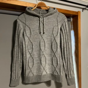 Grå stickad hoodie - Säljer en snygg grå stickad hoodie med kabelstickat mönster. Tröjan har en huva med dragsko och långa ärmar. Perfekt för kyliga dagar!