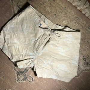 Beige blommiga shorts - Snygga beige shorts med blommigt mönster i ljusblått. Tillverkade i 100% bomull, vilket gör dem bekväma och perfekta för varma dagar. De har en praktisk dragsko i midjan för justerbar passform.