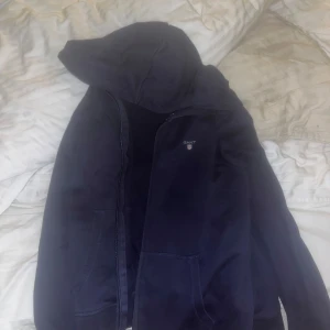 Mörkblå hoodie från Gant - Säljer en mörkblå hoodie från Gant med dragkedja och huva. Perfekt för en avslappnad stil. Den har en liten logotyp på bröstet och är tillverkad i ett mjukt material.