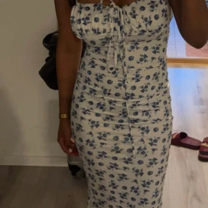 Blommig klänning med knyt - Säljer en söt blommig klänning med blå blommor på vit bakgrund. Klänningen har en knytning framtill och är ärmlös, vilket gör den perfekt för varma dagar. Den har en tight passform som framhäver figuren.