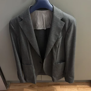 Grå kavaj från Oscar Jacobson - Elegant grå kavaj från Oscar Jacobson med klassisk design. Kavajen har två knappar framtill och fickor på sidorna. Perfekt för en stilren look.