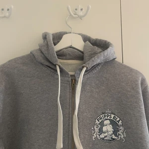 Grå hoodie med Pripps Blå-logga - Grå hoodie med dragkedja och Pripps Blå-logga. Sparsamt använd men som ny.