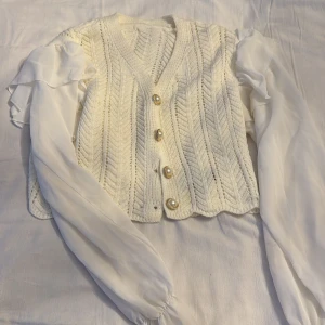 Vit stickad väst med pärlknappar - Säljer en elegant vit stickad väst med v-ringning och dekorativa pärlknappar. Västen har ett fint mönster och långa, transparenta ärmar som ger en unik look. Perfekt för att lägga till en touch av stil till din outfit.