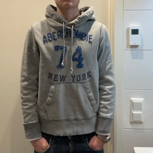 Grå hoodie från Abercrombie - Säljer en grå hoodie från Abercrombie med blå text och siffror på framsidan. skick 10/10 då den nästan inte är använd! slitningarna är design. han på bilden är 183 cm 70 kg. två små prickar vid ena armbågen som knapt syns vid användning