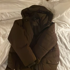 Vero Moda Brun dunjacka med huva - Säljer en varm och mysig brun dunjacka med huva. Jackan har en dragkedja framtill och stora fickor med knappar. Perfekt för kalla dagar.ett litet litet håll i en av fickorna 