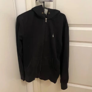 Svart hoodie från Polo Ralph Lauren - Säljer en stilren svart hoodie från Polo Ralph Lauren med dragkedja och huva. Tröjan har en liten logotyp på bröstet och praktiska fickor framtill. Perfekt för en avslappnad look.