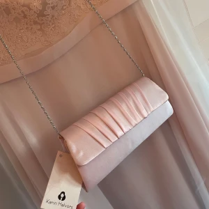 Ljusrosa kuvertväska i satin från Karin Halvors - Elegant ljusrosa kuvertväska i satin med kedjerem från Karin Halvors. Väskan har en stilren design med veckade detaljer på framsidan och stängs med en tryckknapp. Perfekt för speciella tillfällen.