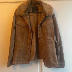 Brun jacka med pälsdetaljer från Tom Tailor - Säljer en snygg brun jacka från Tom Tailor med mjuk pälsdetalj på kragen. Jackan har knappar framtill och två bröstfickor med lock. Perfekt för kyligare dagar.