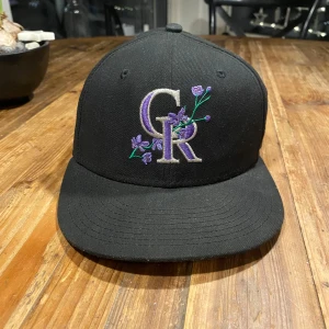 Svart New Era keps med broderad logotyp - Snygg svart keps från New Era med broderad logotyp i lila och silver. Kepsen har en platt skärm och är en del av 59FIFTY-serien. Perfekt för sportiga tillfällen eller som en stilren accessoar.