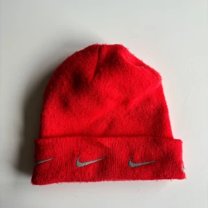 Röd mössa från Nike - Snygg röd mössa från Nike med broderade logotyper i grått. Perfekt för att hålla värmen under kyliga dagar. Den är tillverkad i ett mjukt och bekvämt material.