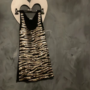Svart och guld glittrig klänning från Quiz - Snygg svart klänning från Quiz med glittriga guldmönster i zebraliknande stil. Perfekt för en kväll ute med vänner. Klänningen är ärmlös och har en rak passform.