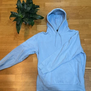 Ljusblå hoodie - Säljer en stilren ljusblå hoodie med justerbar huva och långa ärmar. Perfekt för en avslappnad look. Den har en klassisk design med en stor ficka framtill.