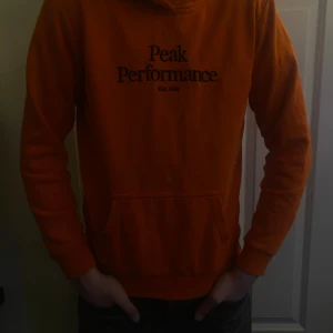 Orange hoodie från Peak Performance - Säljer en snygg orange hoodie från Peak Performance med tryck framtill. Perfekt för en avslappnad stil. Den har en klassisk passform och är tillverkad i mjukt material för extra komfort.