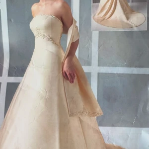 Champagnefärgad axelbandslös bröllopsklänning - Elegant axelbandslös klänning i champagnefärg med vackra broderade detaljer och en svepande lång kjol. Klänningen har en klassisk siluett med knappar längs ryggen och en subtil släp. Perfekt för bröllop och speciella tillfällen.