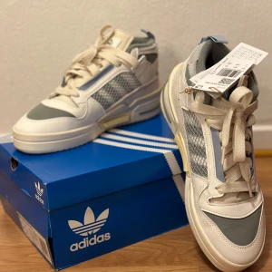 Adidas Forum Mod Mid - Snygga Adidas Forum Mod Mid sneakers i vitt och grått med snörning. Skorna har en modern design med meshdetaljer och en bekväm sula. Perfekta för en stilren look.