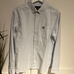 Randig skjorta från Lyle & Scott - Snygg och stilren randig skjorta från Lyle & Scott i ljusblått och vitt. Skjortan har långa ärmar och knappar framtill samt en broderad logotyp på bröstet. Perfekt för en klassisk look.