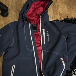 Marinblå jacka från Sail Racing - Snygg marinblå jacka från Sail Racing med röd insida och vita dragkedjor. Jackan har en justerbar huva och flera praktiska fickor. Perfekt för blåsiga dagar.