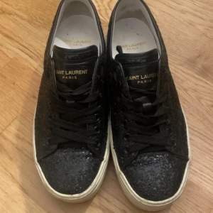 Svarta sneakers från Saint Laurent - Snygga svarta sneakers från Saint Laurent med glittrig yta och klassisk snörning. Skorna har en vit sula och märkeslogga i guld på plösen. Skorna är i storlek 37 och det är anledningen till varför jag säljer dem då jag vanligtvis har 38 eller 39.