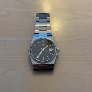 Tissot PRX Automatisk Herrklocka - Snygg Tissot PRX herrklocka med automatisk urverk. Klockan har en stilren svart urtavla med datumvisning och silverfärgade visare. Armbandet är i rostfritt stål med en klassisk länkdesign. Perfekt för den som gillar en tidlös och elegant stil.
