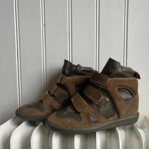 Grönbruna skor från Isabel Marant - Snygga bruna sneakers från Isabel Marant med en unik design. Skorna har en kombination av mocka och skinn med kardborreband för enkel justering. Perfekta för en stilren look. Modellen buckee. Säljer pga fel storlek