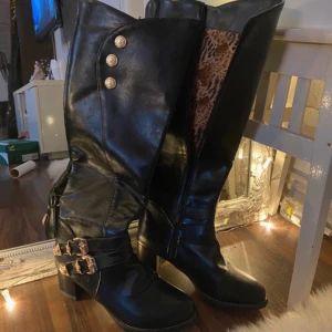 Svarta stövlar med leopardmönstrad insida - Snygga svarta stövlar i skinn med leopardmönstrad insida. De har dekorativa guldfärgade spännen och knappar samt en dragkedja på sidan. Perfekta för en trendig look. Och helt nya aldrig använd 