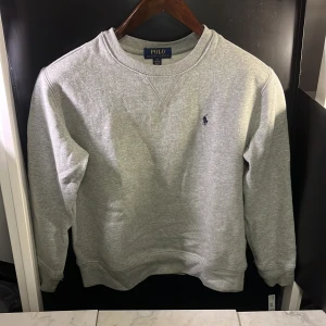 Grå sweatshirt från Polo Ralph Lauren - Säljer en klassisk grå sweatshirt från Polo Ralph Lauren med rund hals och broderad logga på bröstet. Perfekt för en avslappnad stil. Tröjan har långa ärmar och ribbade muddar vid ärmslut och nederkant. Hör av er vid minsta fundering🙌