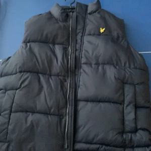 Mörkblå dunväst med dragkedja - Snygg mörkblå dunväst med dragkedja och ett litet gult broderat emblem på bröstet. Perfekt för kyligare dagar och enkel att matcha med olika outfits. Helt ny