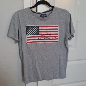 Grå t-shirt från Vinson Polo Club - Säljer en grå t-shirt från Vinson Polo Club med ett tryck av den amerikanska flaggan och texten 'Legendary Vinson Polo Club'. Perfekt för en avslappnad stil. Kan sänka priset vid snabbt köp!
