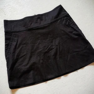 Svart kortkjol med fickor - Snygg svart kortkjol med praktiska fickor. Ett perfekt Basic plagg att ha i sin garderob för en stilren look eller fest. Kjolen har en enkel design som gör den lätt att matcha med olika toppar. Den är i storlek 34 och knappt använd.