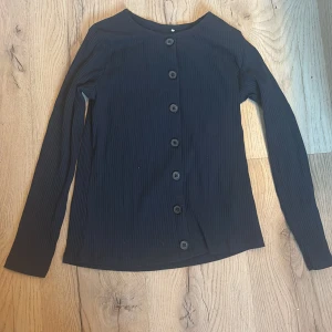 Svart ribbad kofta från Name It - Säljer en stilren svart ribbad kofta från Name It, perfekt för barn i åldern 9-10 år. Koftan har långa ärmar och knappar framtill, tillverkad i ekologisk bomull för extra komfort. Ett mångsidigt plagg för alla tillfällen!