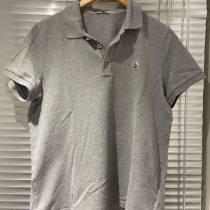  pikétröja från Moncler - Snygg grå pikétröja från Moncler med korta ärmar och klassisk krage. Tröjan har en broderad logotyp på bröstet och knappar i halsen med röd och vit detalj. Den är i storlek L men sitter som M.