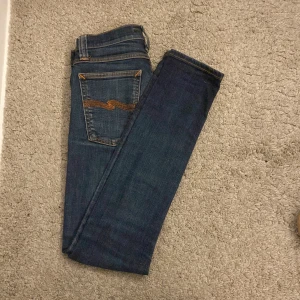 Blå jeans från Nudie Jeans - Snygga blå jeans från Nudie Jeans med klassisk femficksdesign och orange sömmar. Perfekta för en avslappnad stil. De har en knappgylf och är tillverkade i högkvalitativ denim.