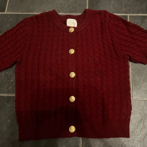 Vinröd stickad kofta med guldknappar - Säljer en vinröd stickad kofta med guldknappar. Koftan är kortärmad. Använd en gång