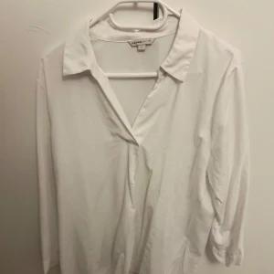Vit blus  - Elegant vit blus med en klassisk krage och v-ringning. Blusen har trekvartsärmar och är tillverkad i ett mjukt material som ger en stilren look. Perfekt för en chic och minimalistisk stil.