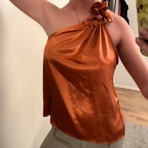 Orange satintopp - Snygg orange satin one-shoulder / halterneck topp med knytning vid axeln. Perfekt för en kväll ute eller en speciell tillställning. Den glansiga ytan ger en lyxig känsla.